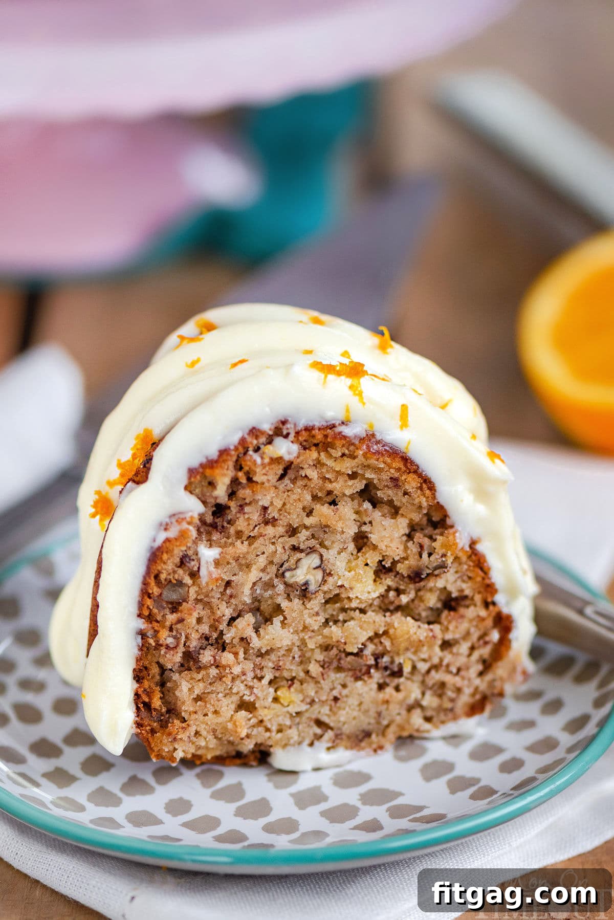 A slice of moist Hummingbird Bundt Cake, generously topped with creamy cream cheese frosting, resting on a small dessert plate in front of an elegant pink cake stand. The cake reveals its delightful banana and pineapple-infused crumb and visible pecan pieces.