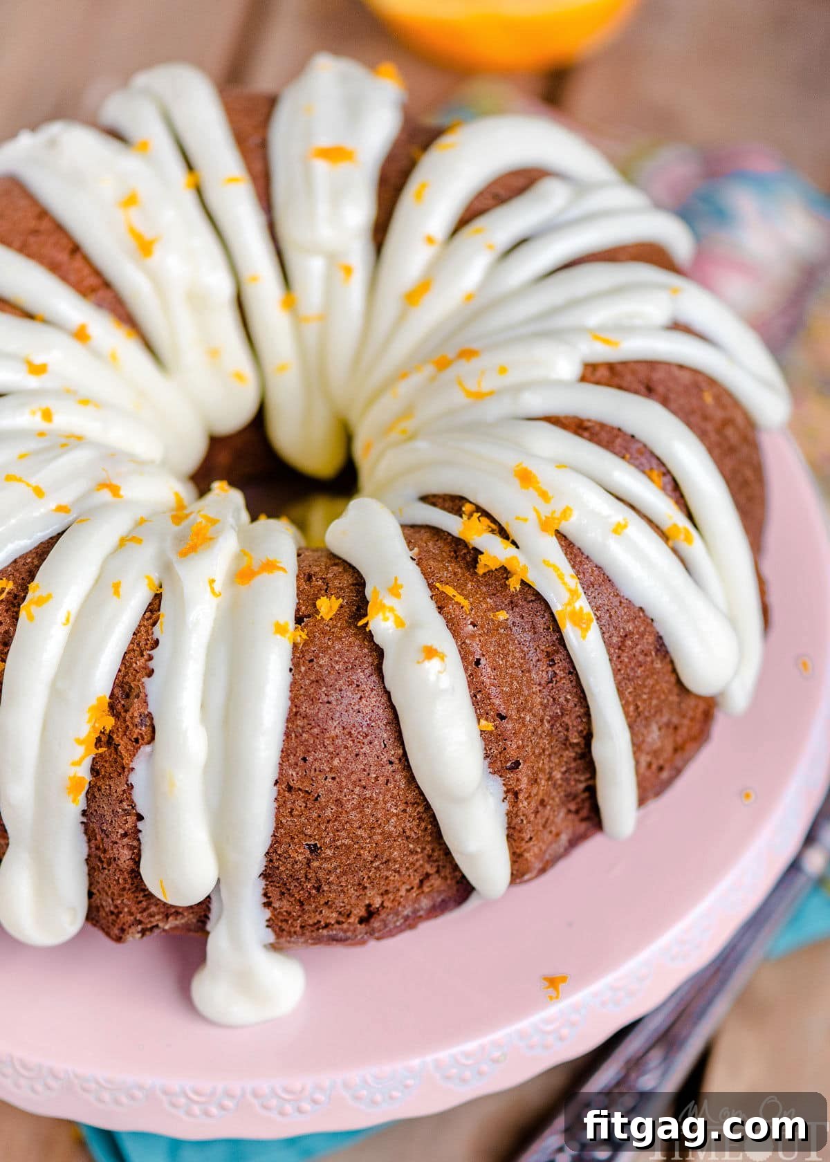 A top-down, angled view of a beautiful Hummingbird Bundt Cake, generously covered with a silky cream cheese frosting and garnished with vibrant orange zest. The intricate design of the bundt pan is highlighted by the glaze.