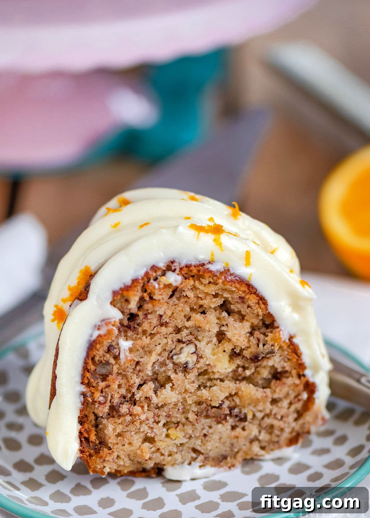 A charming piece of Hummingbird Bundt Cake, generously topped with creamy white frosting, served on a small, elegant dessert plate beside a pink cake stand.