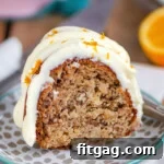 A perfectly sliced piece of Hummingbird Bundt Cake, showcasing its moist texture and topped with a creamy orange-infused cream cheese frosting, rests on a small dessert plate in front of a decorative pink cake stand.
