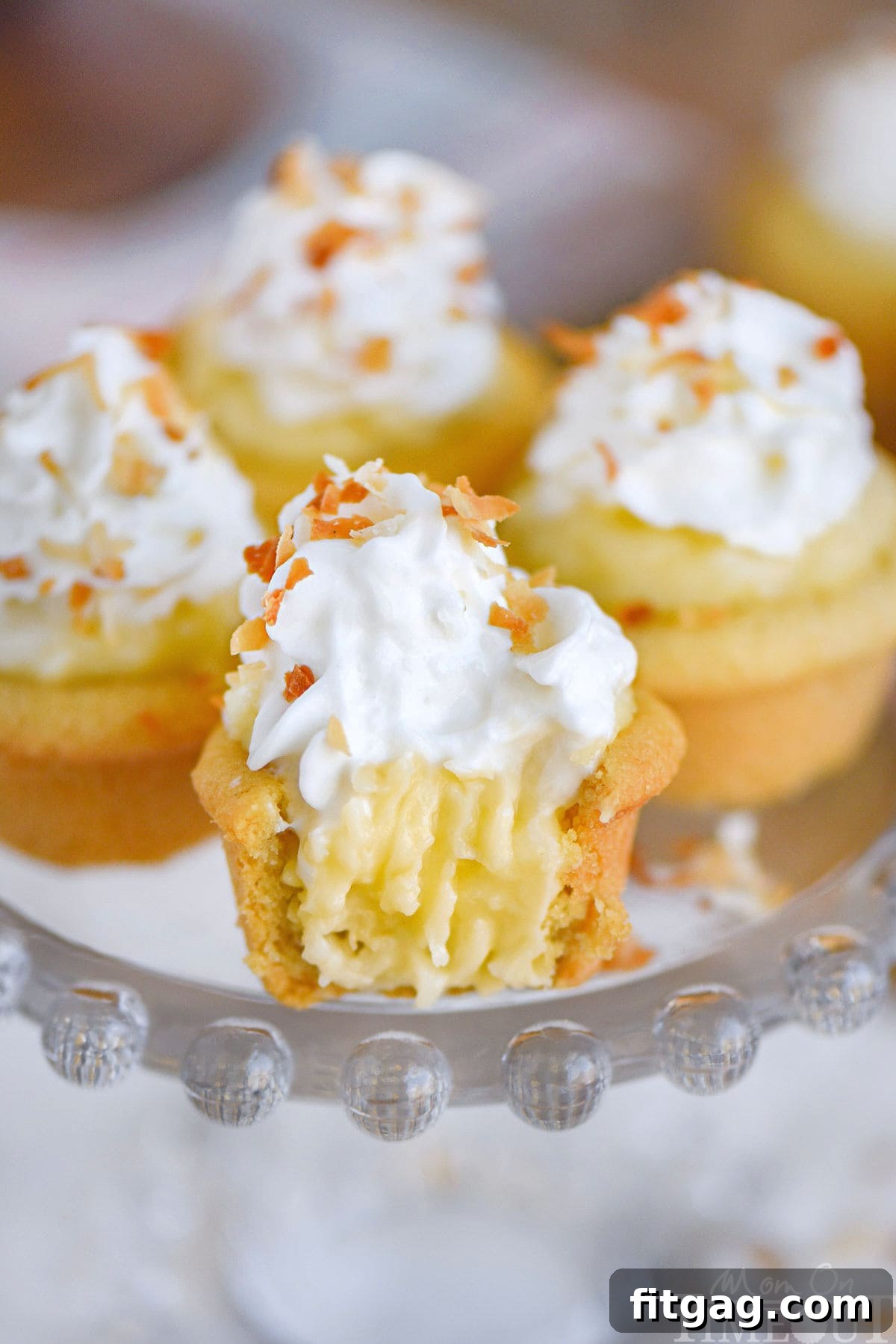 Four mini coconut cream pies on glass cake stand topped with whipped cream and toasted coconut. The front one has a bite taken out of it.