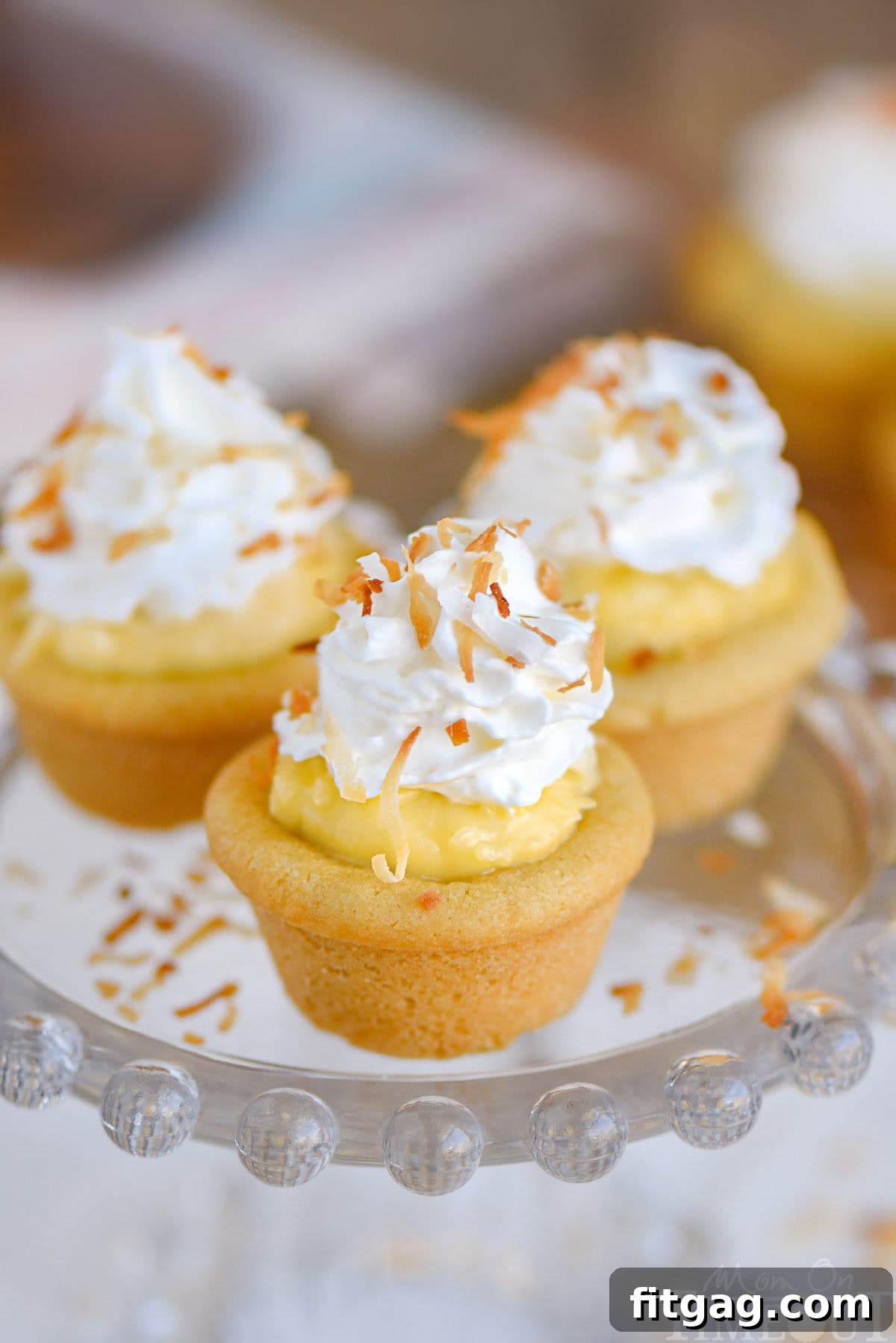 Three coconut cream pie cookies cups on glass stand with whipped cream and toasted coconut on top.