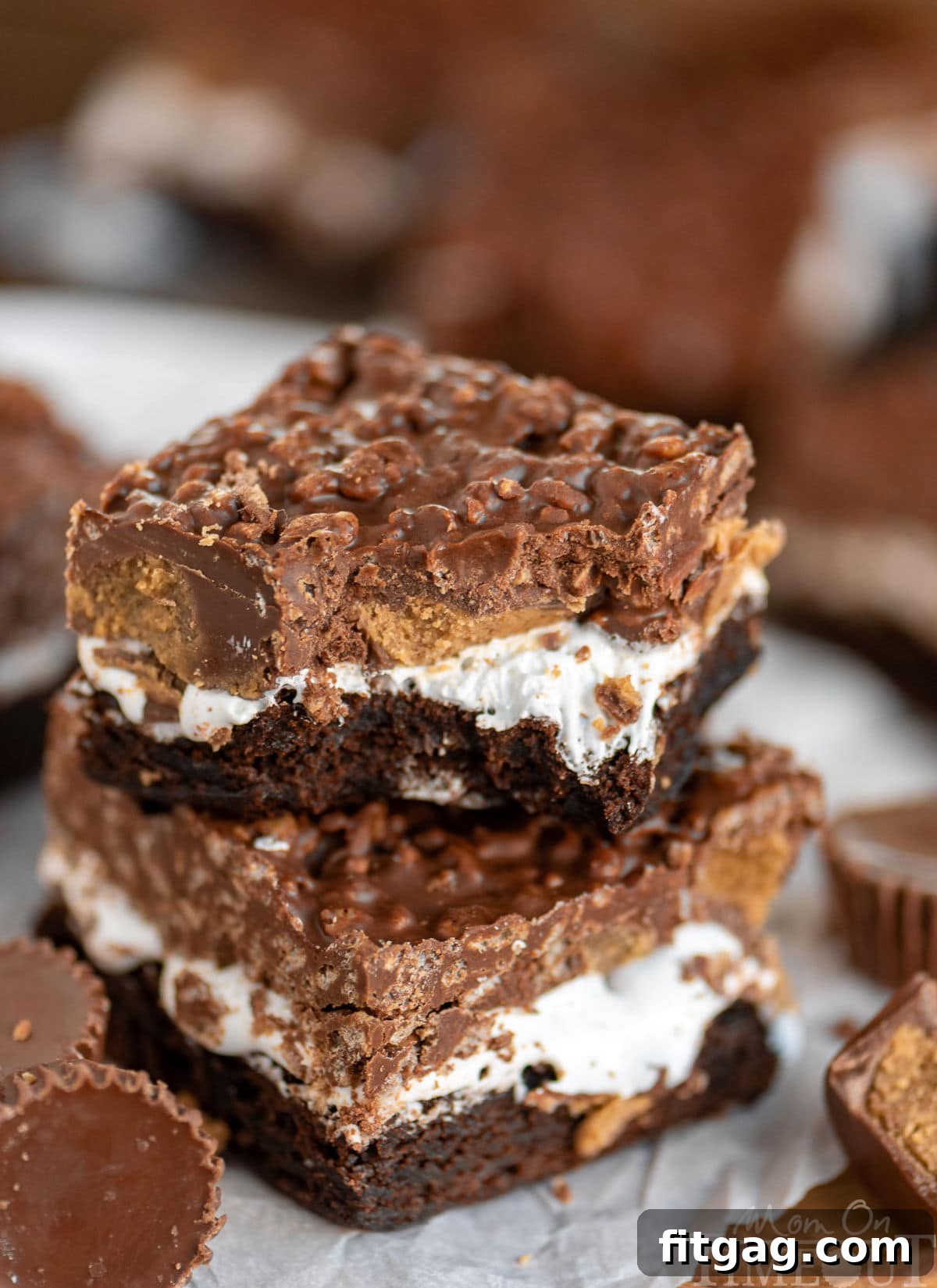 Peanut Butter Cup Marshmallow Brownies 3 Two Reese's Marshmallow Brownies with visible layers, one with a bite taken, and scattered Reese's cups, highlighting the gooey marshmallow and crispy topping.
