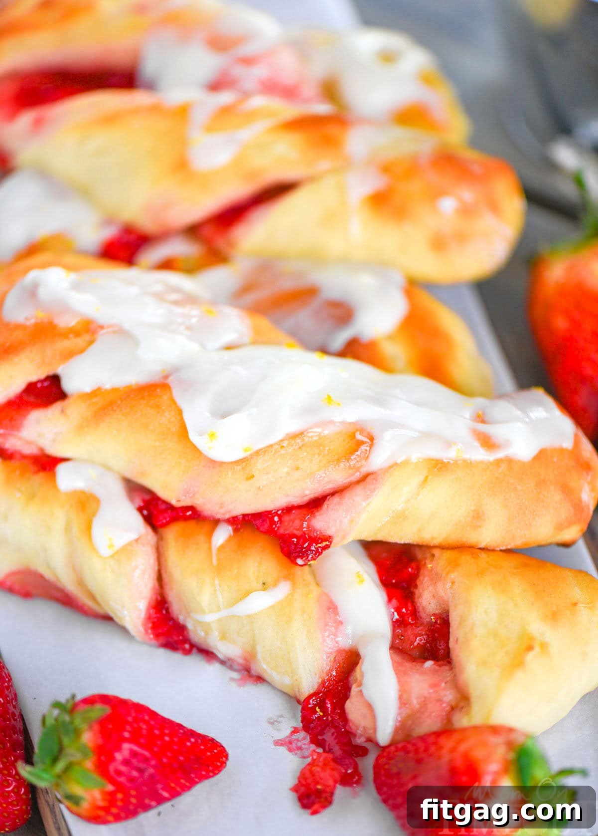 Two golden-brown Strawberry Twists, generously coated with a luscious lemon glaze, are stacked elegantly with fresh strawberries nestled alongside them, highlighting their irresistible appeal.