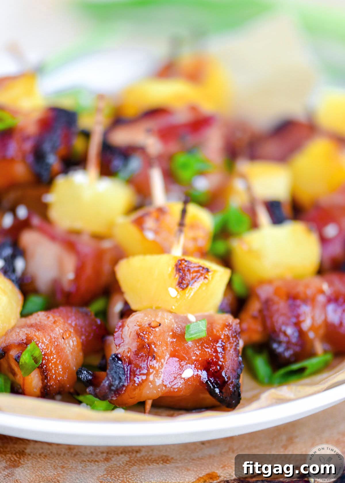 Plate of bacon wrapped chicken teriyaki bites with pineapple. Each bite is skewered with a toothpick and topped with green onions and sesame seeds.