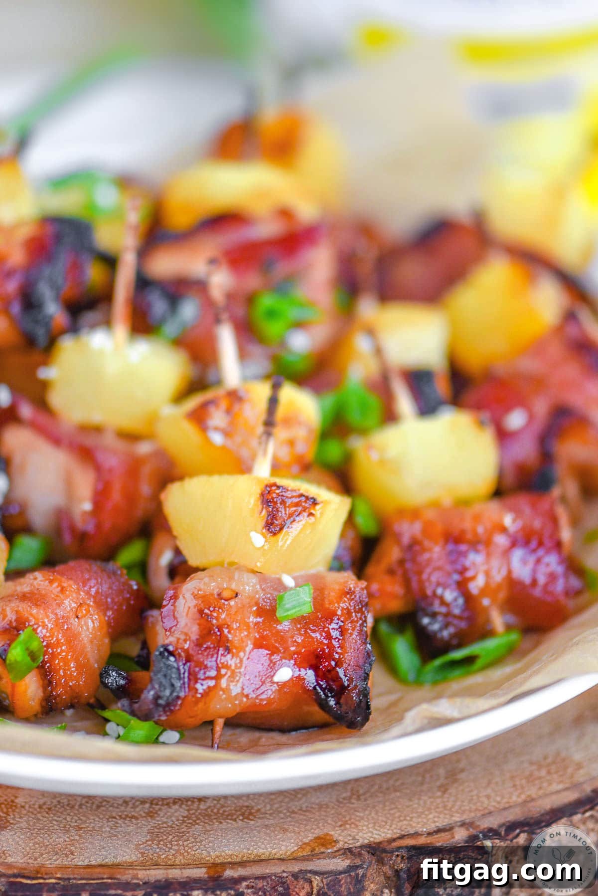 Plate of bacon wrapped chicken teriyaki bites with pineapple. Each bite is skewered with a toothpick and topped with green onions and sesame seeds.