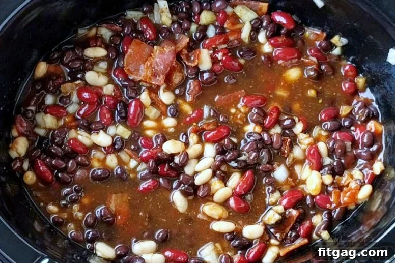 Slow Cooker Kielbasa and Barbecue Beans is the perfect chilly day recipe! Made with three different beans, molasses, bacon, and kielbasa - it's pure comfort food! // Mom On Timeout #slowcooker #crockpot #recipes #bacon #beans #bbq #kielbasa #comfortfood #dinnerideas #recipe Slow Cooker Kielbasa and Barbecue Beans all mixed together in a crockpot, showcasing the rich ingredients.