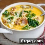 White bowl with handles filled with zuppa toscana soup and topped with cheese. Kale, sausage and bacon can be seen in the soup.