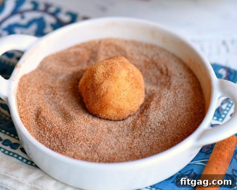Snickerdoodle cookie dough ball being rolled lovingly in a bowl of cinnamon sugar mixture, promising a delightful treat.