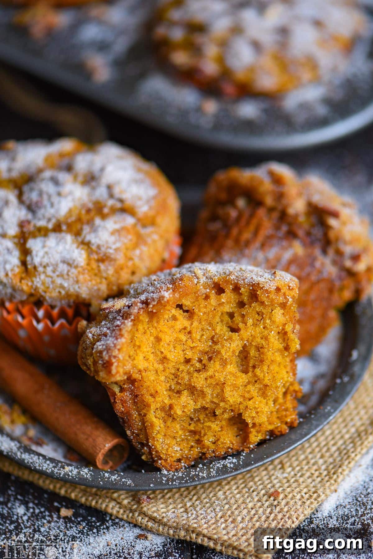 Three perfectly baked pumpkin muffins sitting on a rustic metal plate, accompanied by fragrant cinnamon sticks. The muffins are golden brown with a delightful crackled topping, evoking a cozy fall atmosphere.