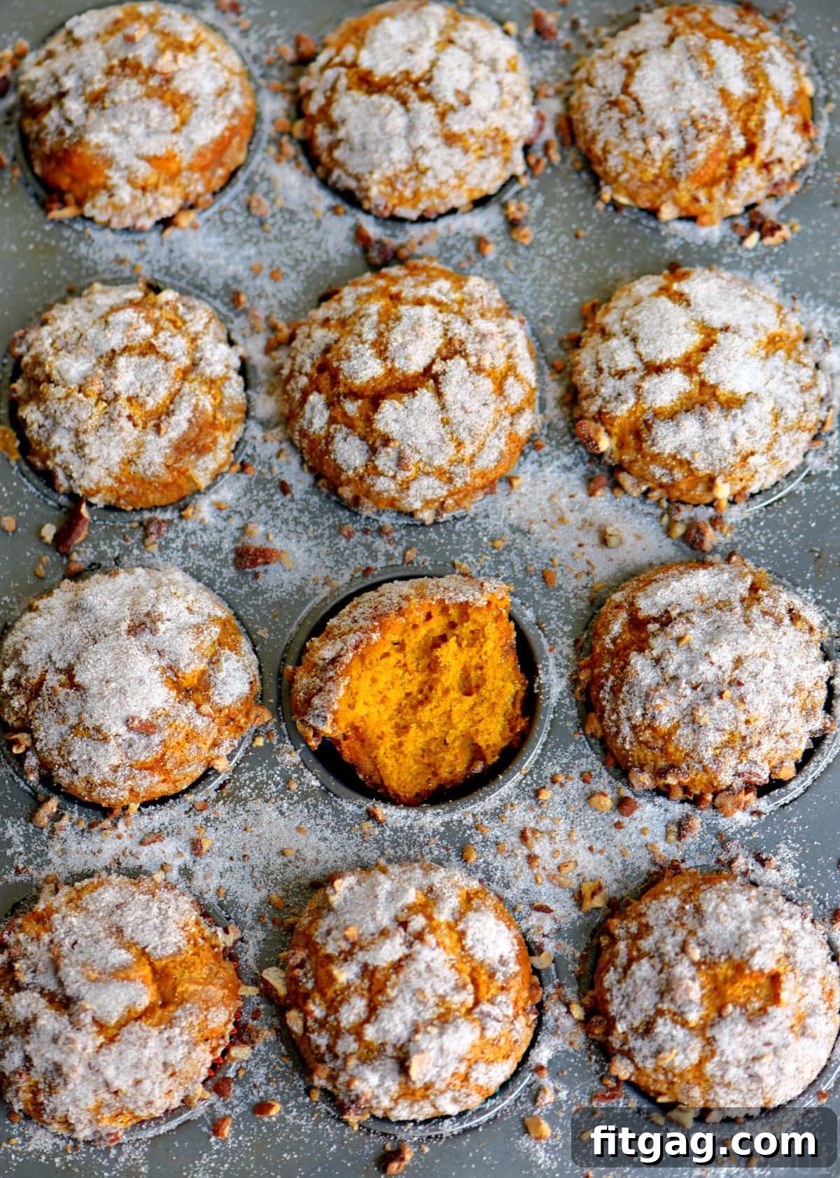 An overhead shot showcasing a batch of freshly baked pumpkin muffins still in the muffin pan, with their signature crackled, sugared tops. One muffin has been thoughtfully torn in half, revealing its incredibly soft, moist, and fluffy interior.