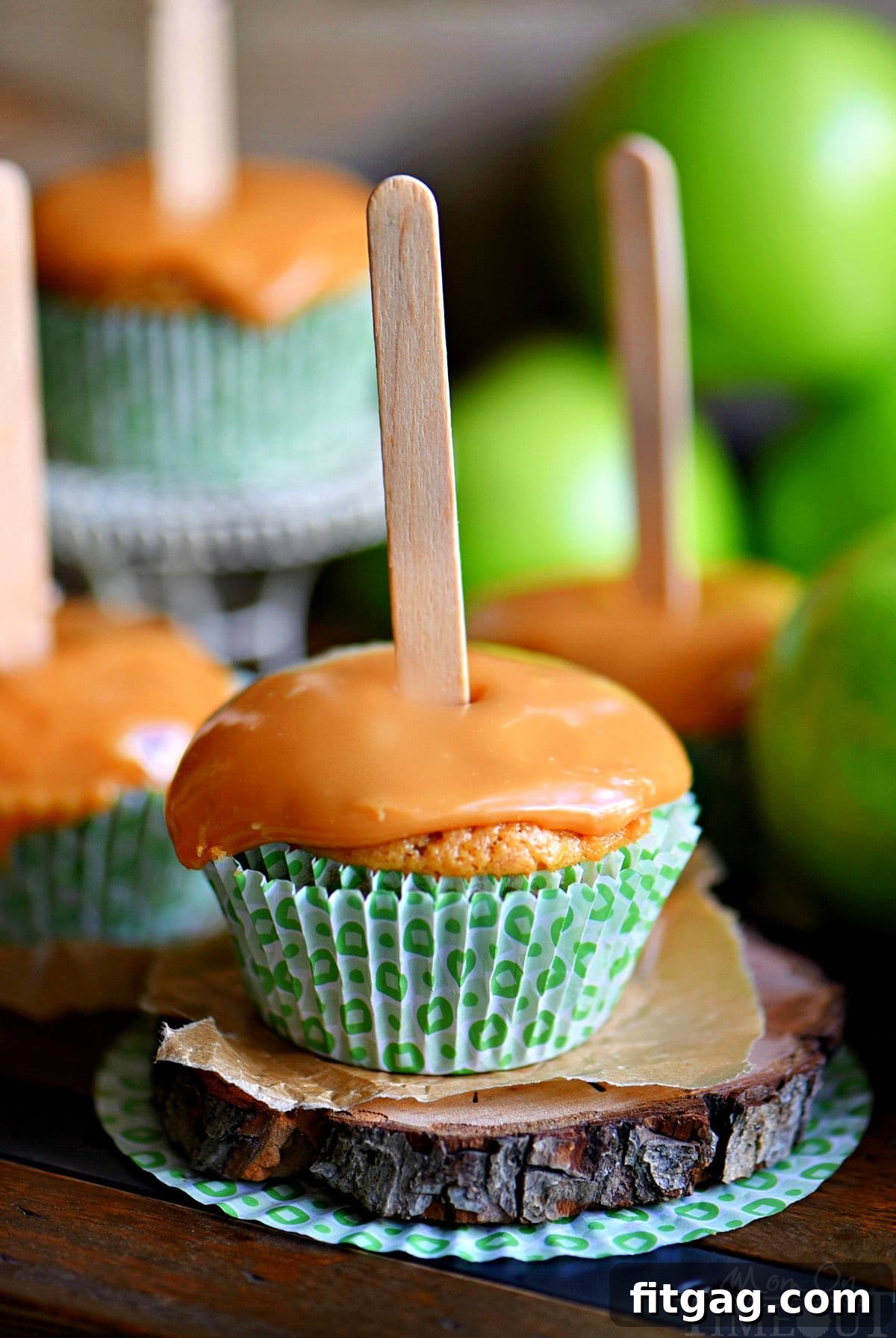 Autumn Caramel Apple Delights 2 Delicious apple cupcake topped with a generous swirl of caramel frosting and a charming popsicle stick inserted into the top. Two more equally tempting cupcakes are visible in the background, set against a warm autumn backdrop.