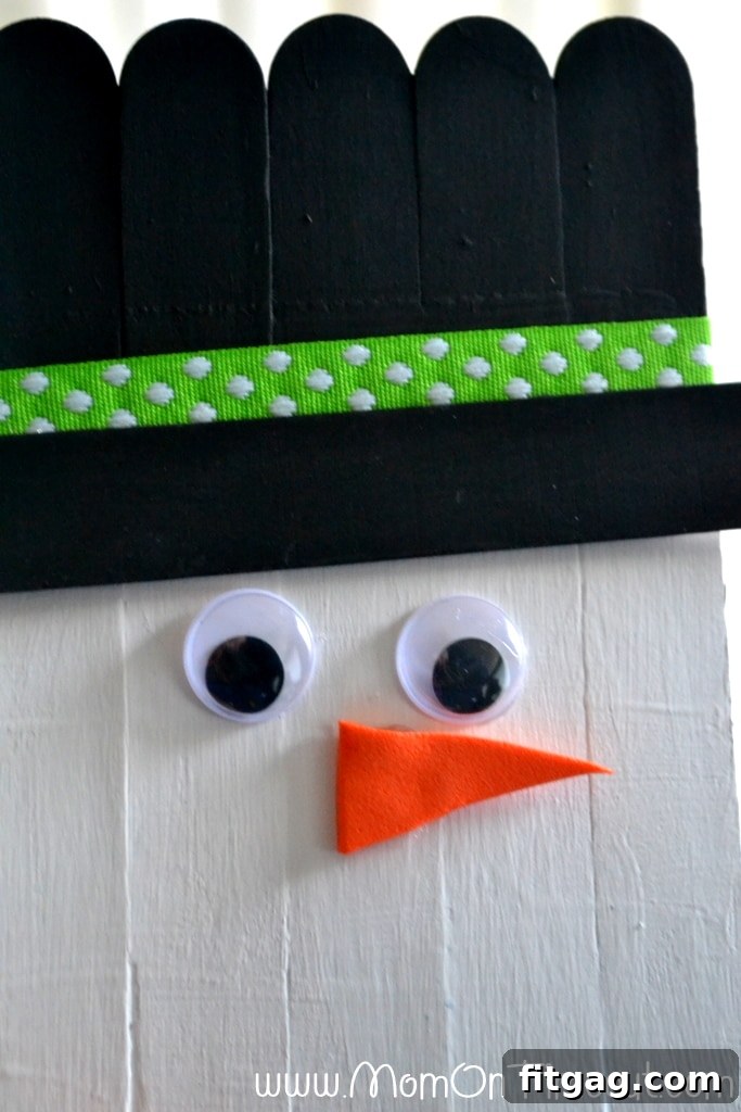 Craft stick snowman with googly eyes, an orange foam nose, and a drawn smile