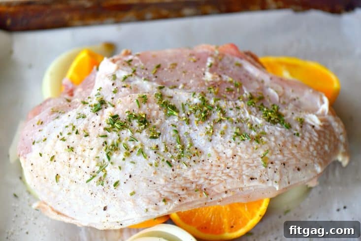 A sheet pan with a turkey breast, surrounded by orange slices, onion wedges, and fresh herbs, ready for the oven.