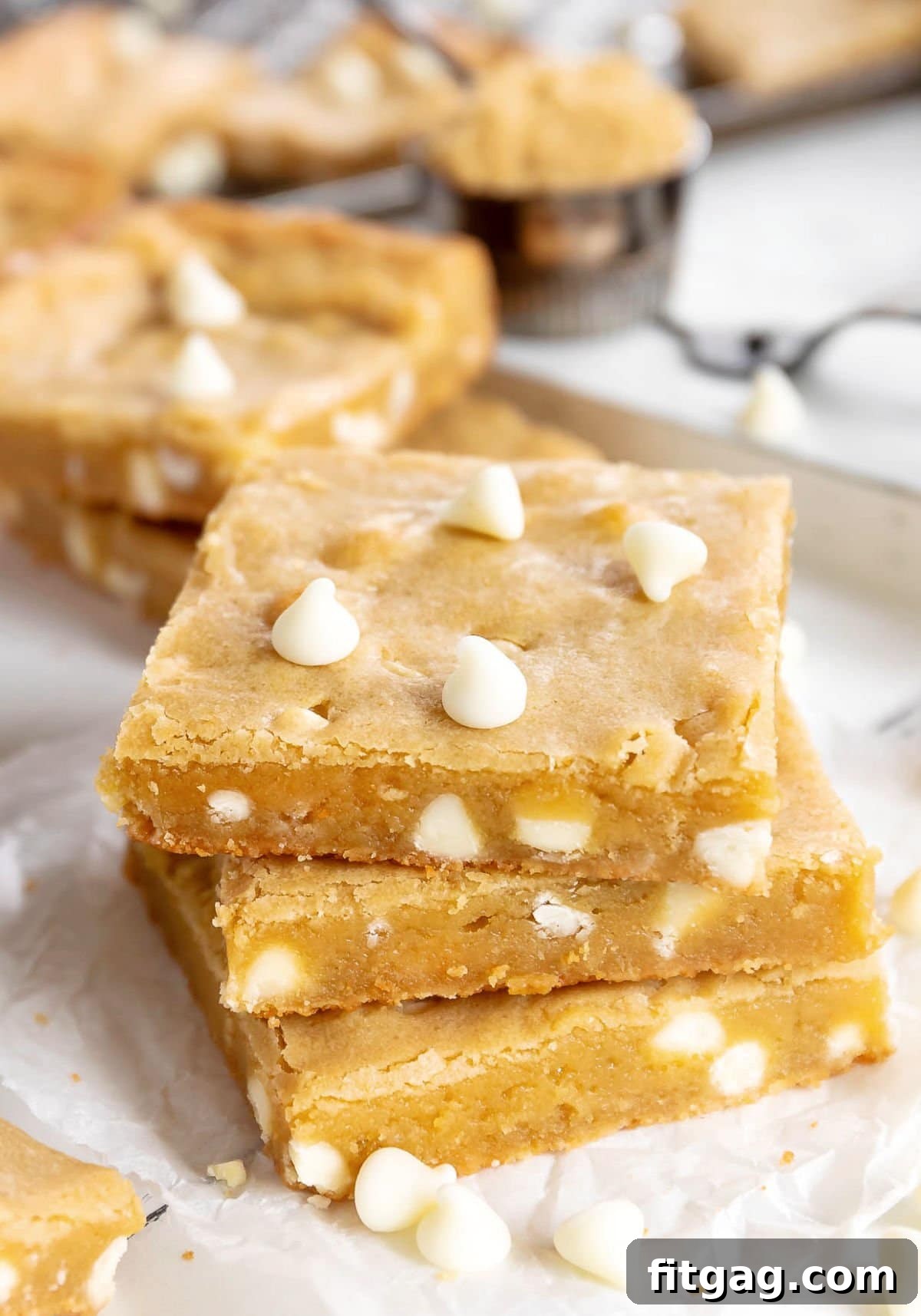Ultimate Blondie Bliss 2 Three perfectly golden blondie bars stacked, adorned with white chocolate chips, resting on white parchment paper. More blondies are visible in the background, hinting at a generous batch of this irresistible dessert.