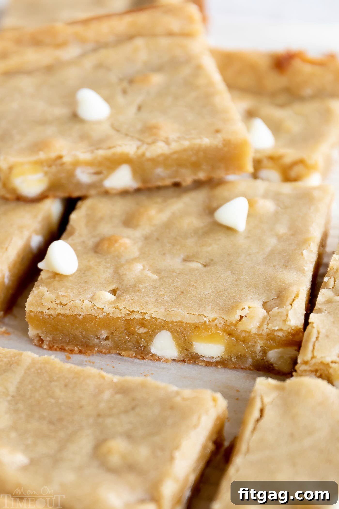 The Best Blondies I've ever had! This easy, one-bowl blondie recipe yields extra chewy, buttery, rich, blondies with hardly any effort at all! Freshly baked blondies, made with creamy white chocolate chips, precisely cut into perfect squares and neatly arranged on pristine white parchment paper. One blondie sits atop the pile, showcasing its golden-brown crust and tempting texture.