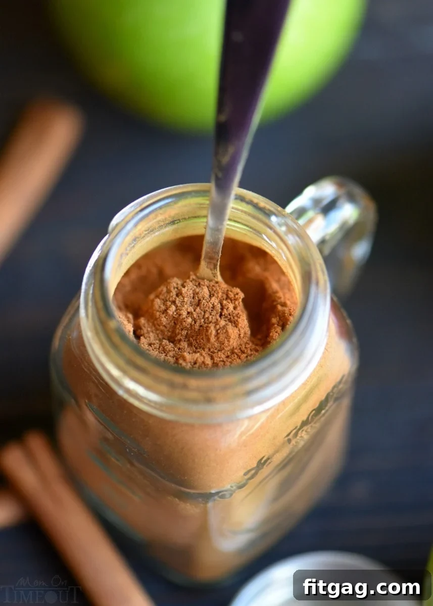 Homemade Apple Pie Spice in Storage Jar A small, clear glass spice jar, filled with homemade apple pie spice, with a miniature wooden spoon resting inside, ready for use. The jar is placed on a light, textured surface.
