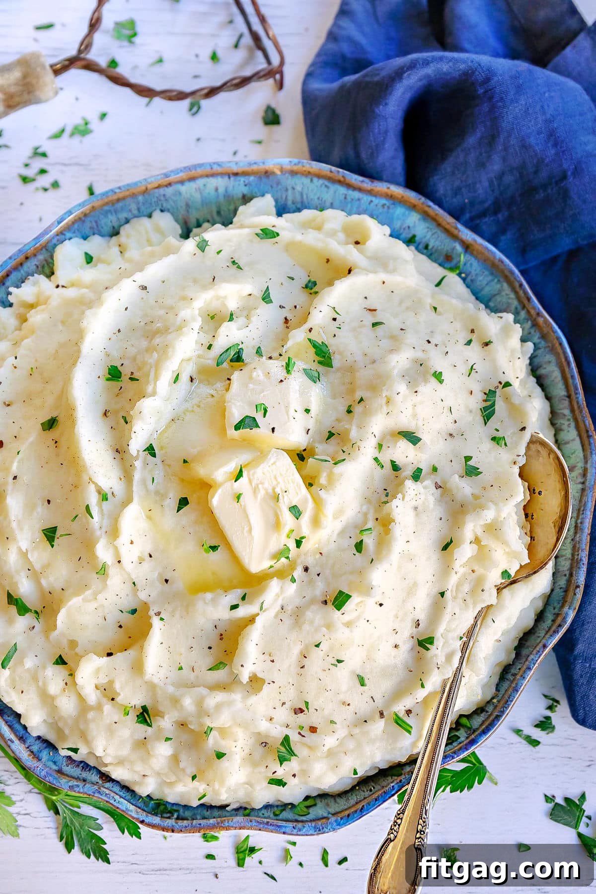 top down look at a blue and green bowl filled with fluffy mashed potatoes. slices of butter, pepper and chopped parsley garnish the mashed potatoes.