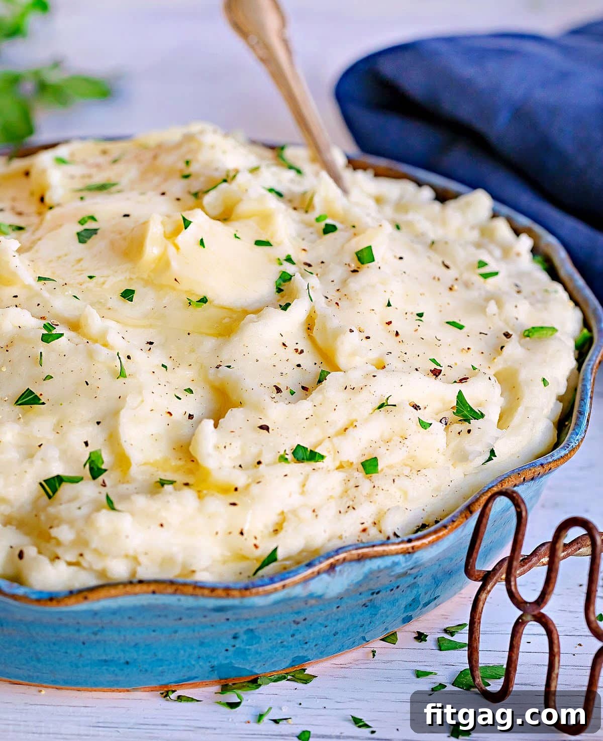 best mashed potatoes in serving bowl topped with thick slices of butter and freshly chopped parsley. blue napkin in background.