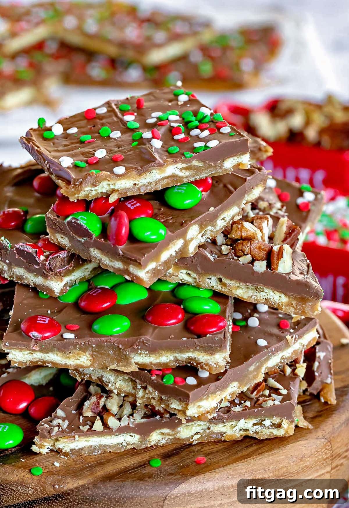Christmas Toffee Craze 2 Various pieces of Christmas Crack, a sweet and salty saltine cracker toffee, piled high on a wooden stand. The toffee is generously topped with colorful M&M's, chopped nuts, and festive sprinkles. More toffee pieces are visible in the background.