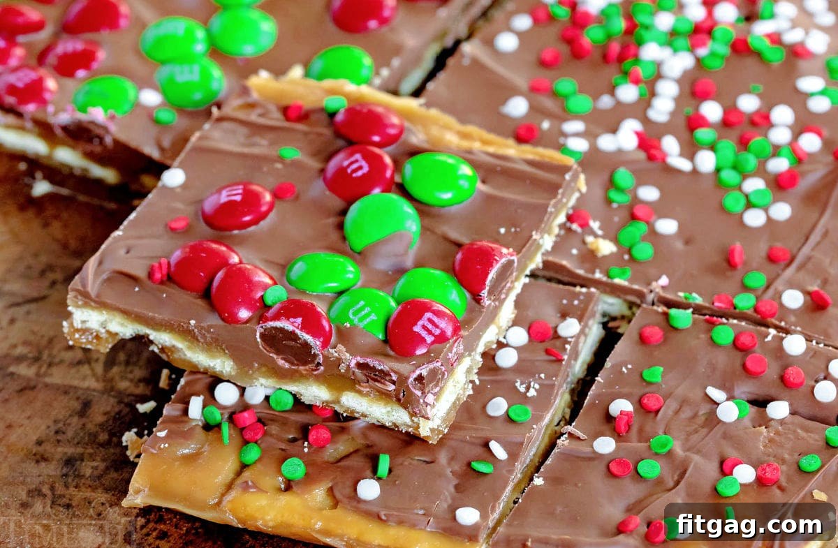 Christmas Toffee Craze 5 A perfectly cut piece of Christmas Crack, generously topped with colorful M&M's, rests on a larger sheet of saltine cracker toffee that is still awaiting its final cut into individual servings. The layers of cracker, caramel, chocolate, and toppings are clearly visible.