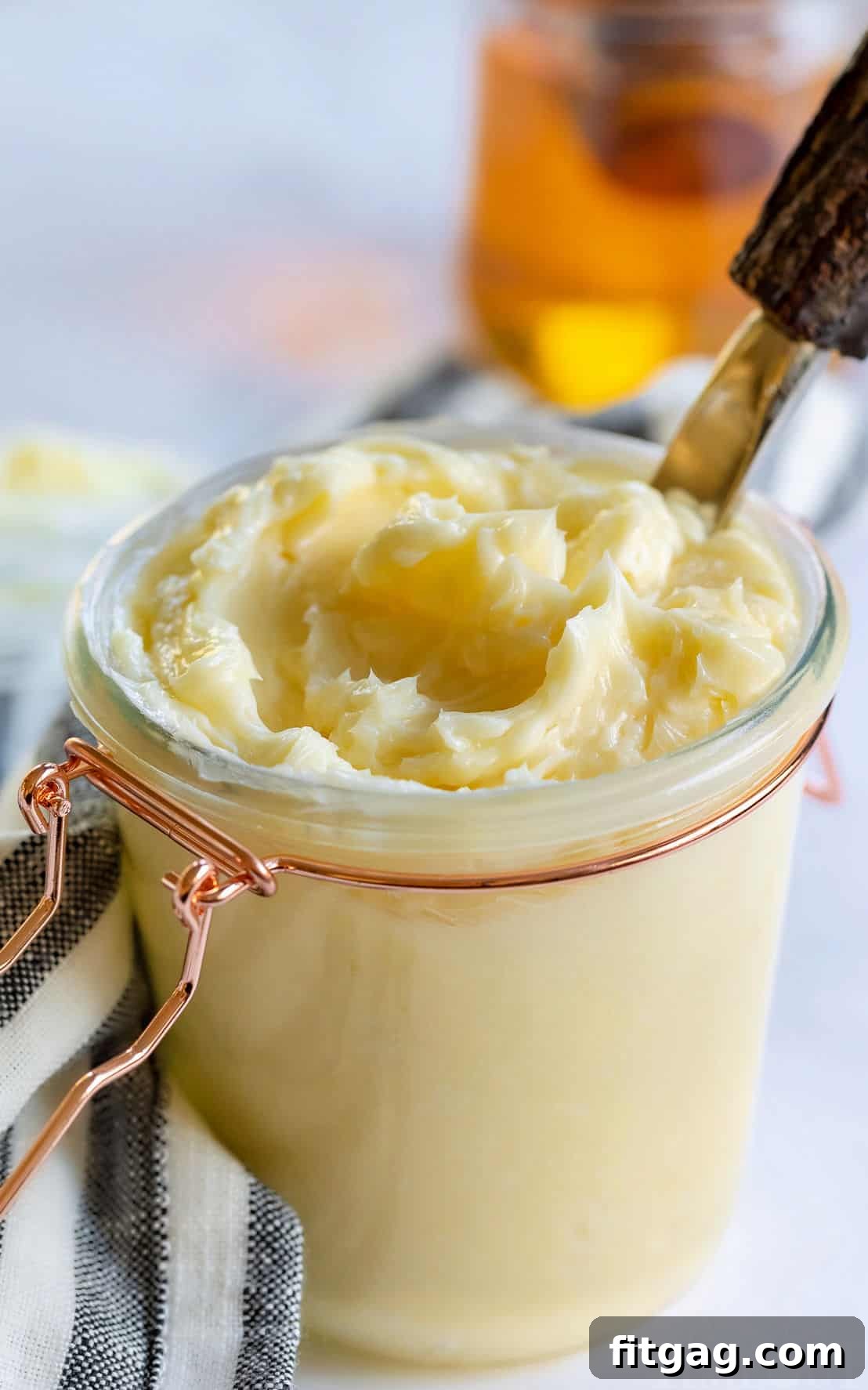 The BEST Honey Butter is ridiculously easy to make and tastes amazing! With just 4 ingredients, this homemade honey butter recipe is perfectly sweet and creamy and is incredible smeared on cornbread, biscuits and homemade dinner rolls! // Mom On Timeout #honeybutter #honeybutterrecipe #homemade #condiments #butter #honey A glass jar filled with creamy honey butter, sealed with a lid and placed on a kitchen surface with a towel, ready for storage or serving.