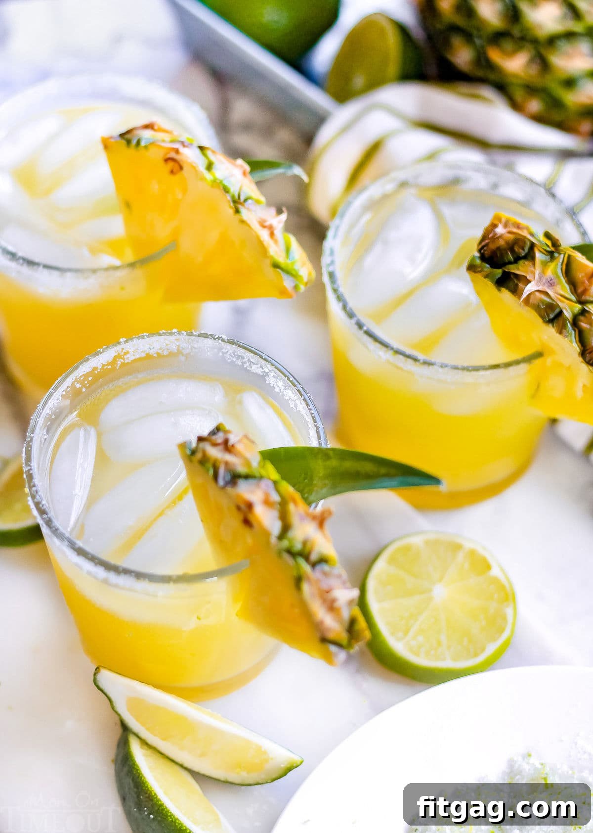 Pineapple Paradise Margarita 3 Three pineapple margaritas on a marble surface with lime slices and pineapple wedges, next to a bowl of lime margarita salt.
