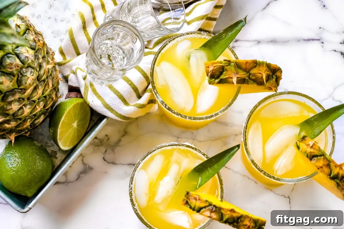 Pineapple Paradise Margarita 4 An easy pineapple margarita recipe displayed in glasses next to a whole pineapple and fresh limes on a pristine white marble backdrop.