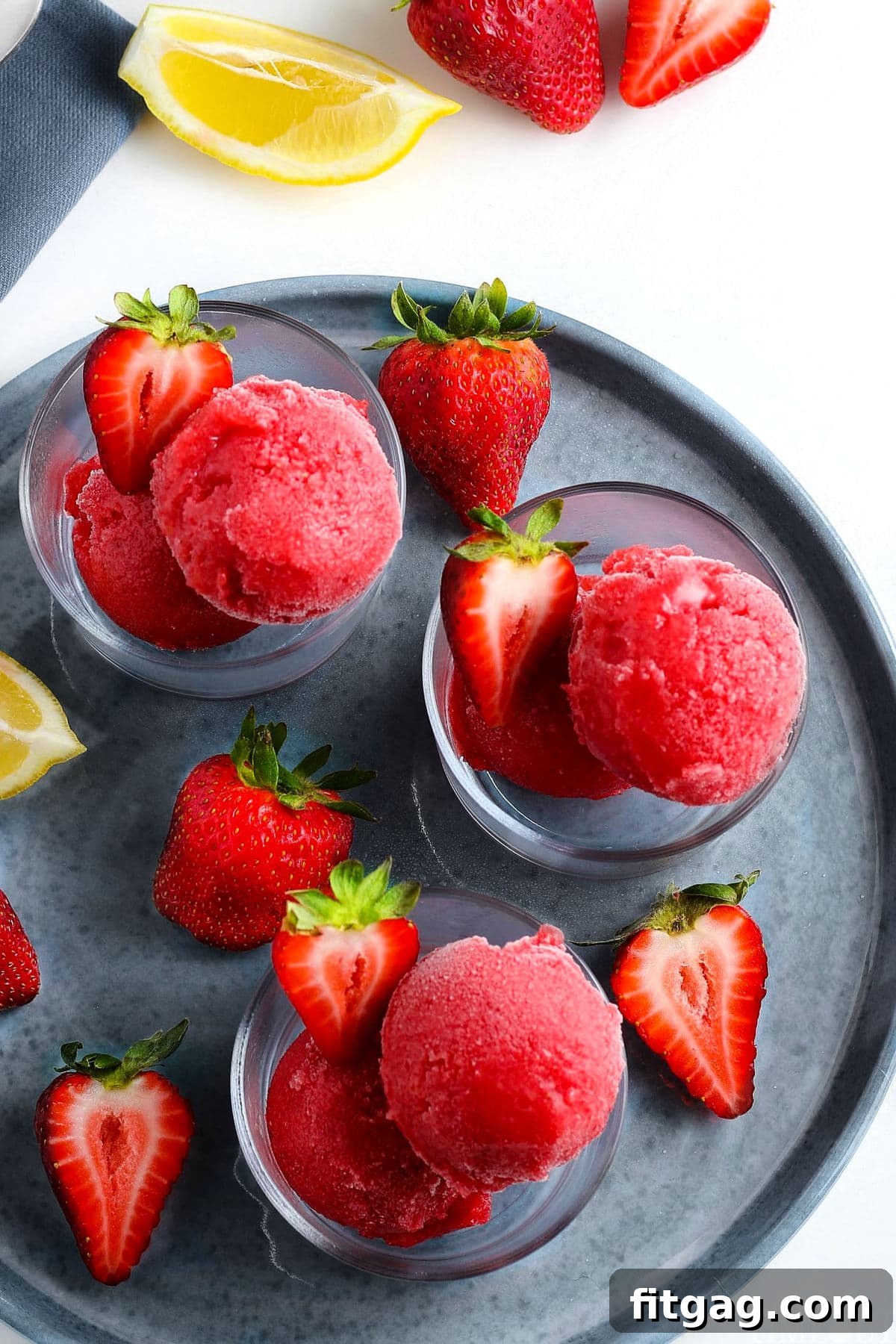 Ruby Frost 3 Three small glasses filled with vibrant strawberry sorbet, garnished with halved fresh strawberries, artfully arranged on a rustic gray plate, highlighting its refreshing appeal.