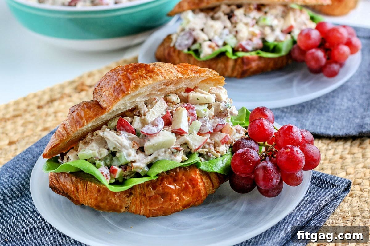 Chicken salad elegantly presented on a croissant, resting on a glass plate with fresh grapes on the side and a blue napkin underneath.