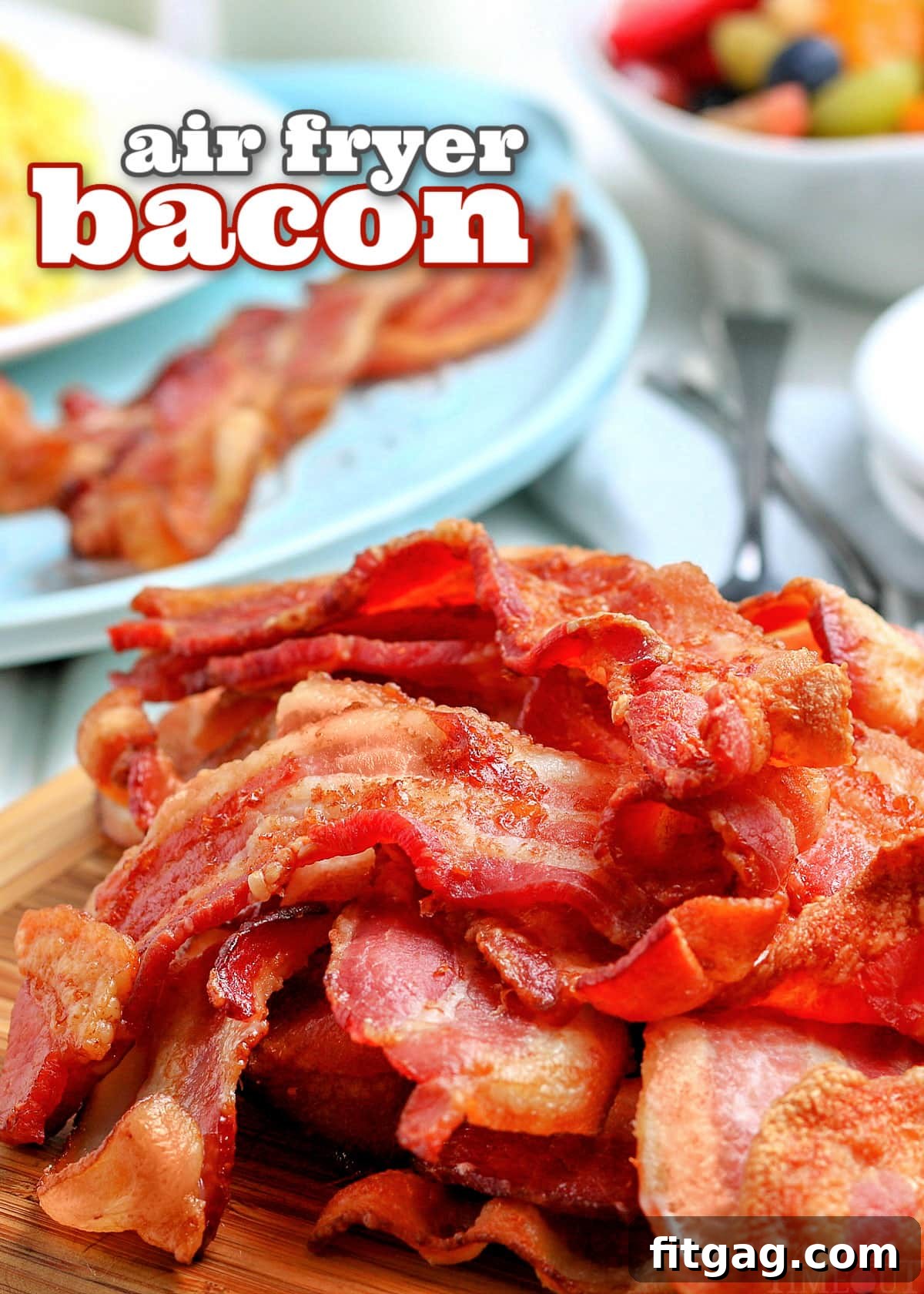 Air fryer bacon piled high on a wooden board with a blue plate of eggs and bacon in the background, along with a bowl of fruit.