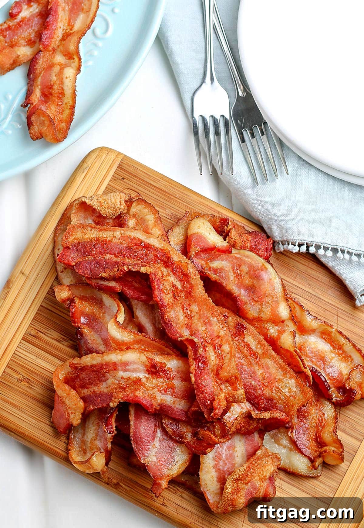 Top-down view of perfectly cooked air fryer bacon piled high on a wooden cutting board, with a stack of white plates nearby.