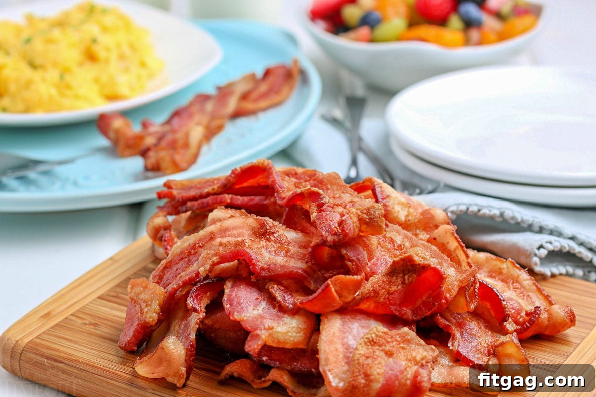 Air fryer bacon piled high on a wooden cutting board with a bowl of fruit salad and a plate with eggs and bacon in the background.