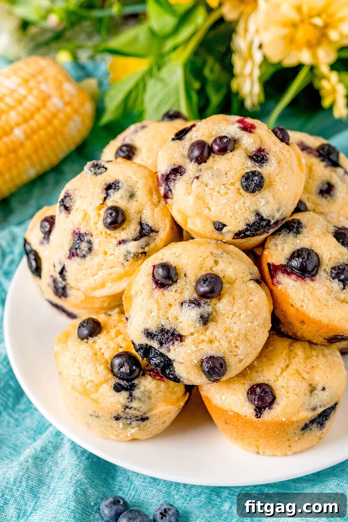 Golden Blueberry Cornbread Muffins 4 Top-down view of blueberry corn muffins on a white plate with corn cobs and yellow flowers in the background