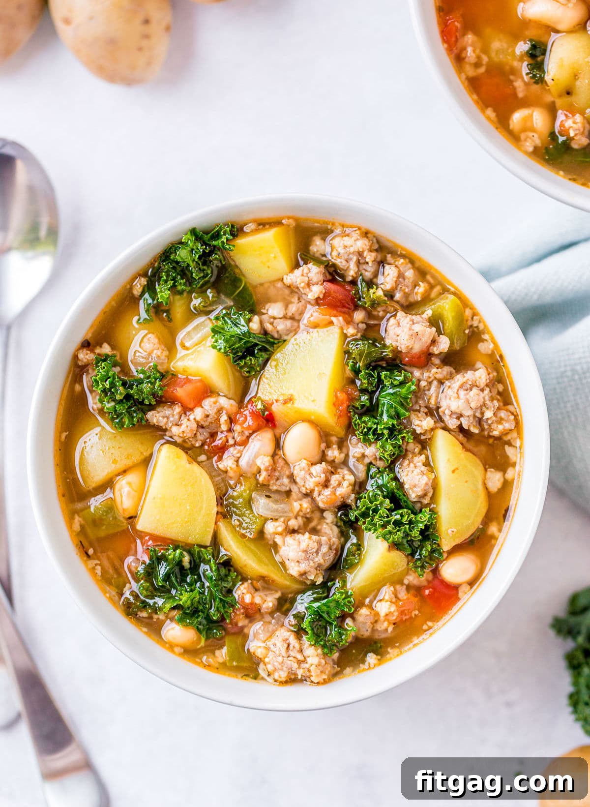 Top-down view of a large serving of sausage potato kale soup with two spoons resting on the side, inviting you to dig in.