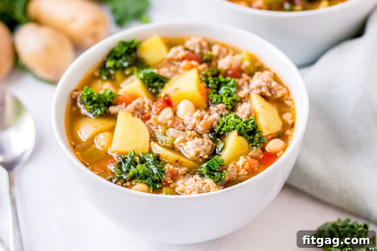 A beautiful bowl of sausage kale soup resting on a white background, accompanied by a spoon and more ingredients in the background.