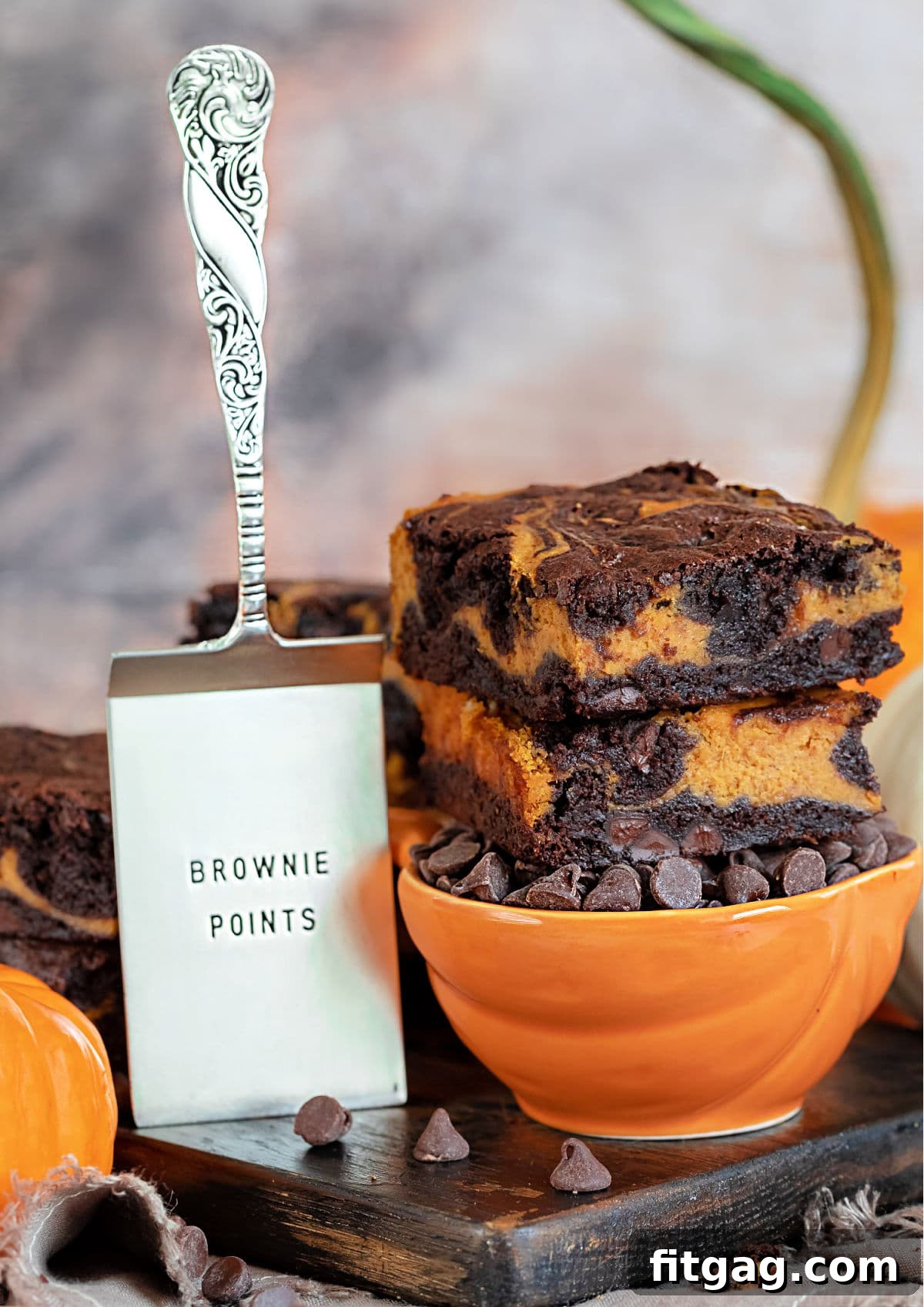 Pumpkin cheesecake brownies sitting on an orange bowl filled with chocolate chips, with a brownie server on the left side.