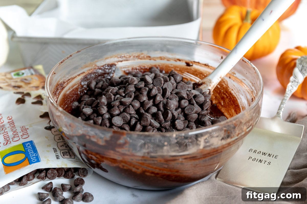 Chocolate chips on top of brownie batter in a glass bowl with chocolate chips scattered about and a baking pan in the background.