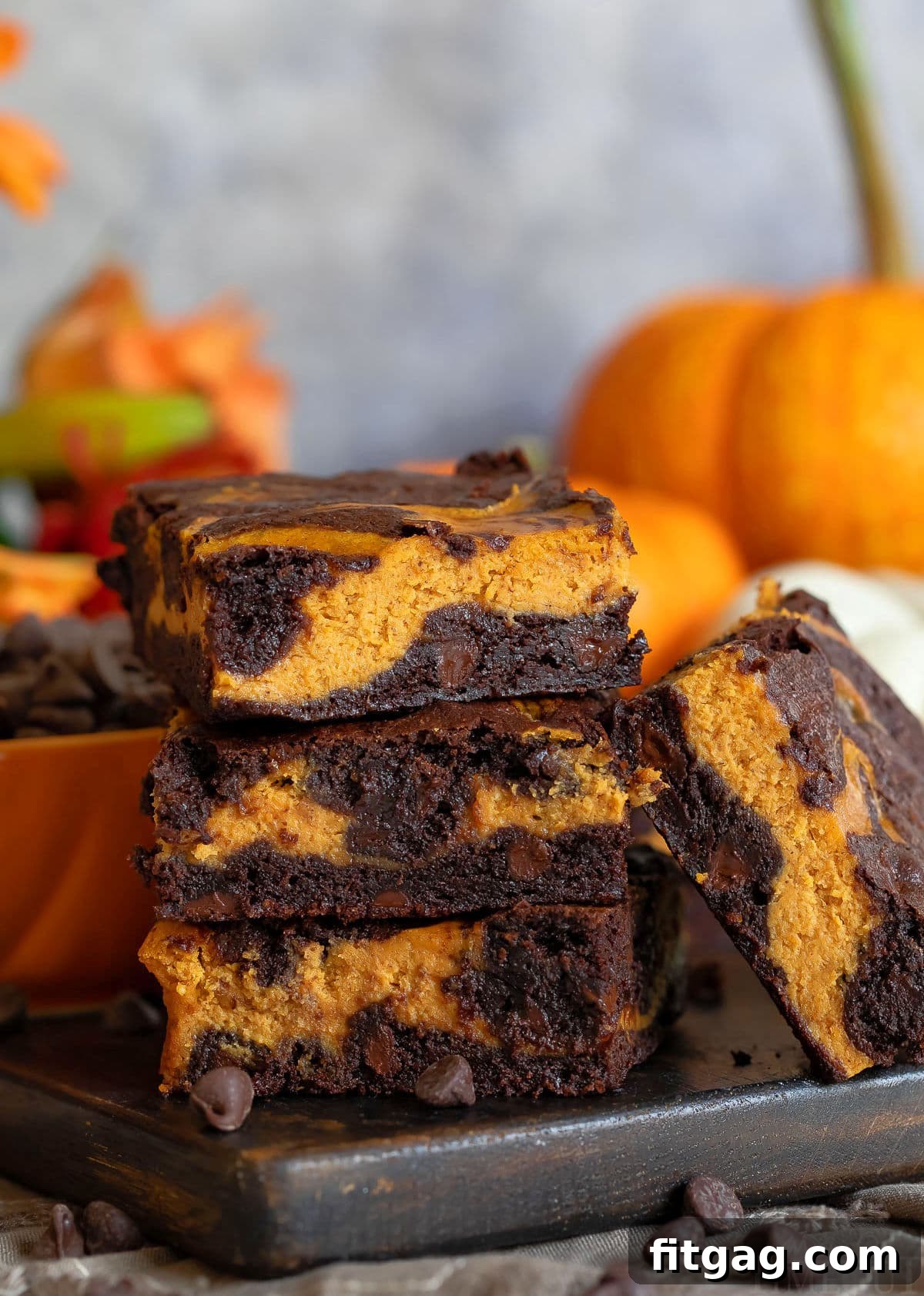 Pumpkin brownies stacked three high on a dark wood board with a pumpkin in the background.