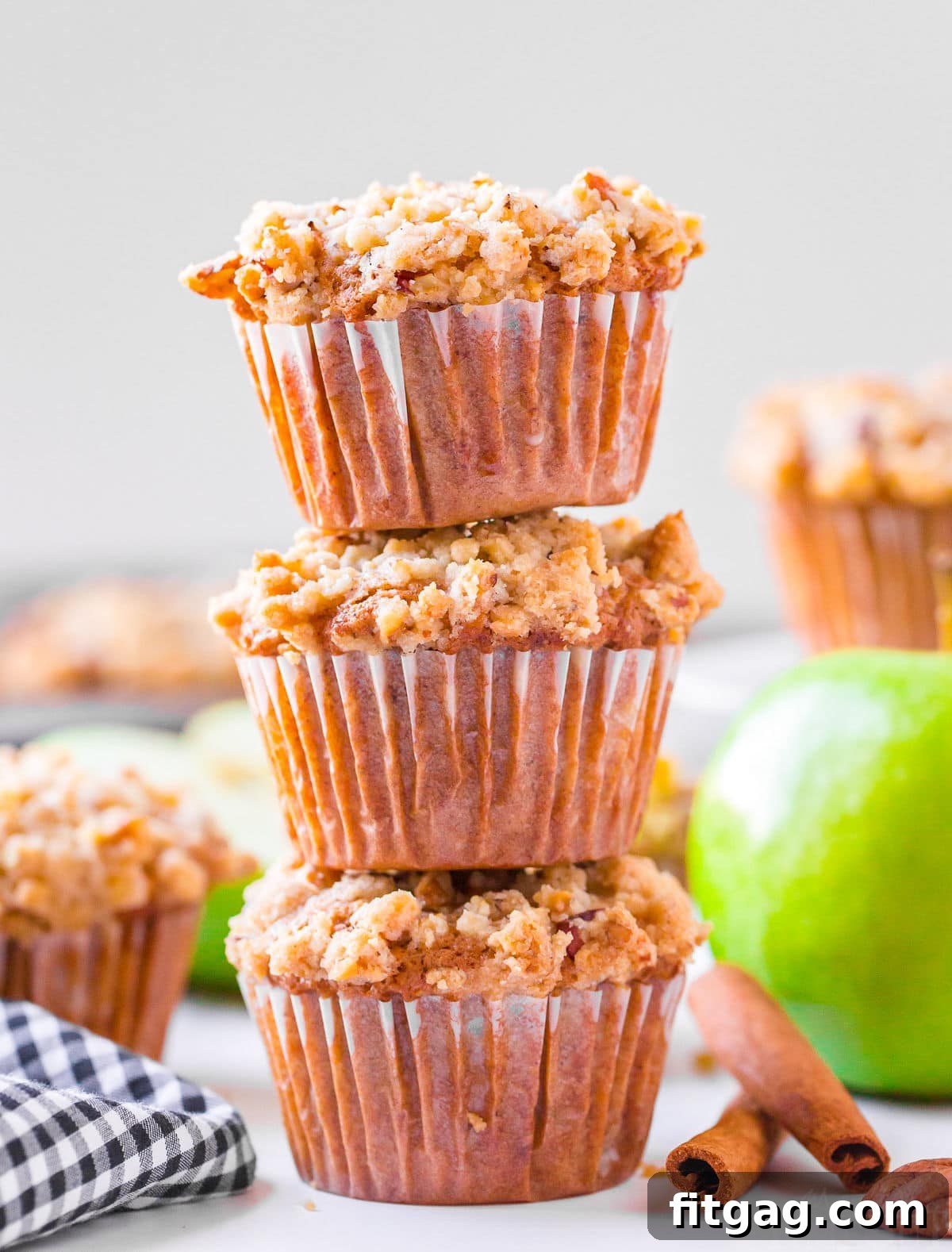 Three golden-brown apple crumb muffins stacked high on a rustic surface, with more delicious muffins softly blurred in the background.
