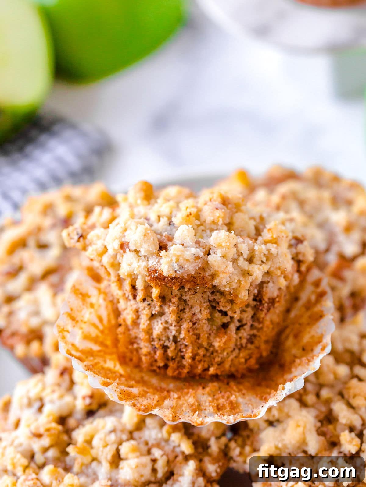 An unwrapped apple muffin with a beautiful pecan crumb topping, resting on a plate with more muffins in the background, ready to be enjoyed.