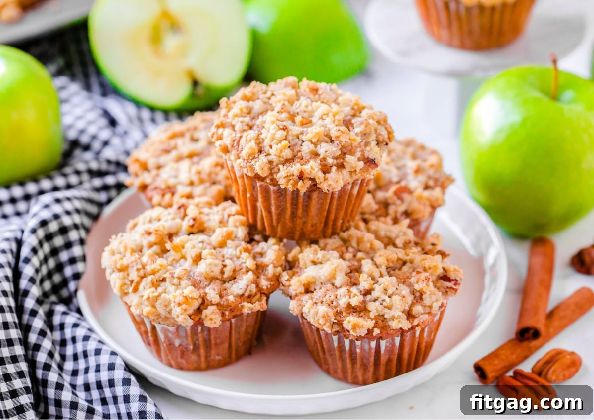 A stack of perfectly golden apple muffins on a white plate, with a blue and white striped napkin peeking out, and fresh apples blurred in the background, hinting at the key ingredient.