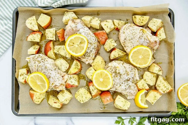 Uncooked Greek chicken and diced baby red potatoes spread out on a parchment-lined sheet pan, marinated and ready for baking in the oven.