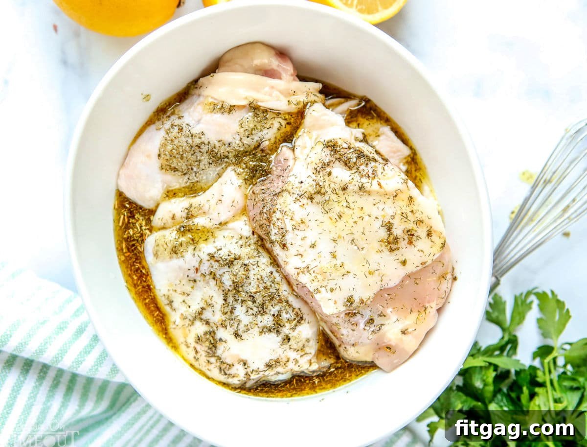 Succulent chicken thighs coated in a rich Greek marinade, resting in a white bowl. Fresh lemons and vibrant parsley are artfully placed nearby, hinting at the dish's fresh flavors.