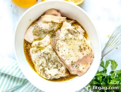 Raw chicken thighs in a white bowl, generously coated with green Greek marinade. Fresh lemons and parsley are nearby on a striped towel.