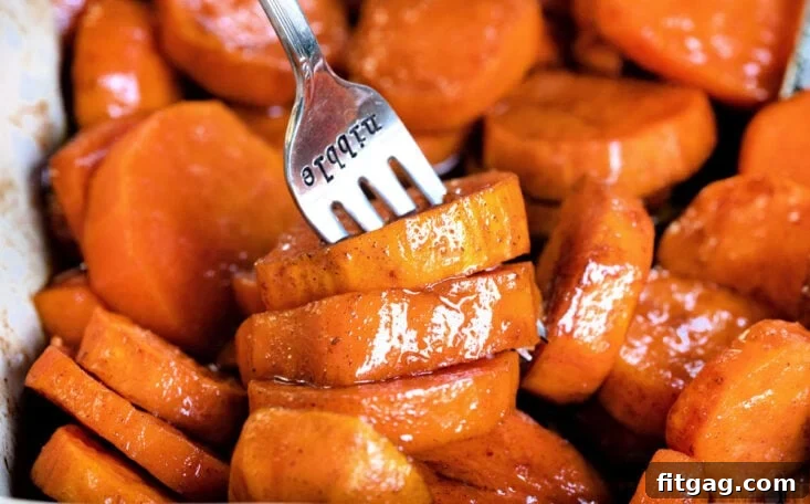 A fork is gently lifting a piece of tender, candied sweet potato from a beige baking dish, highlighting its soft texture.