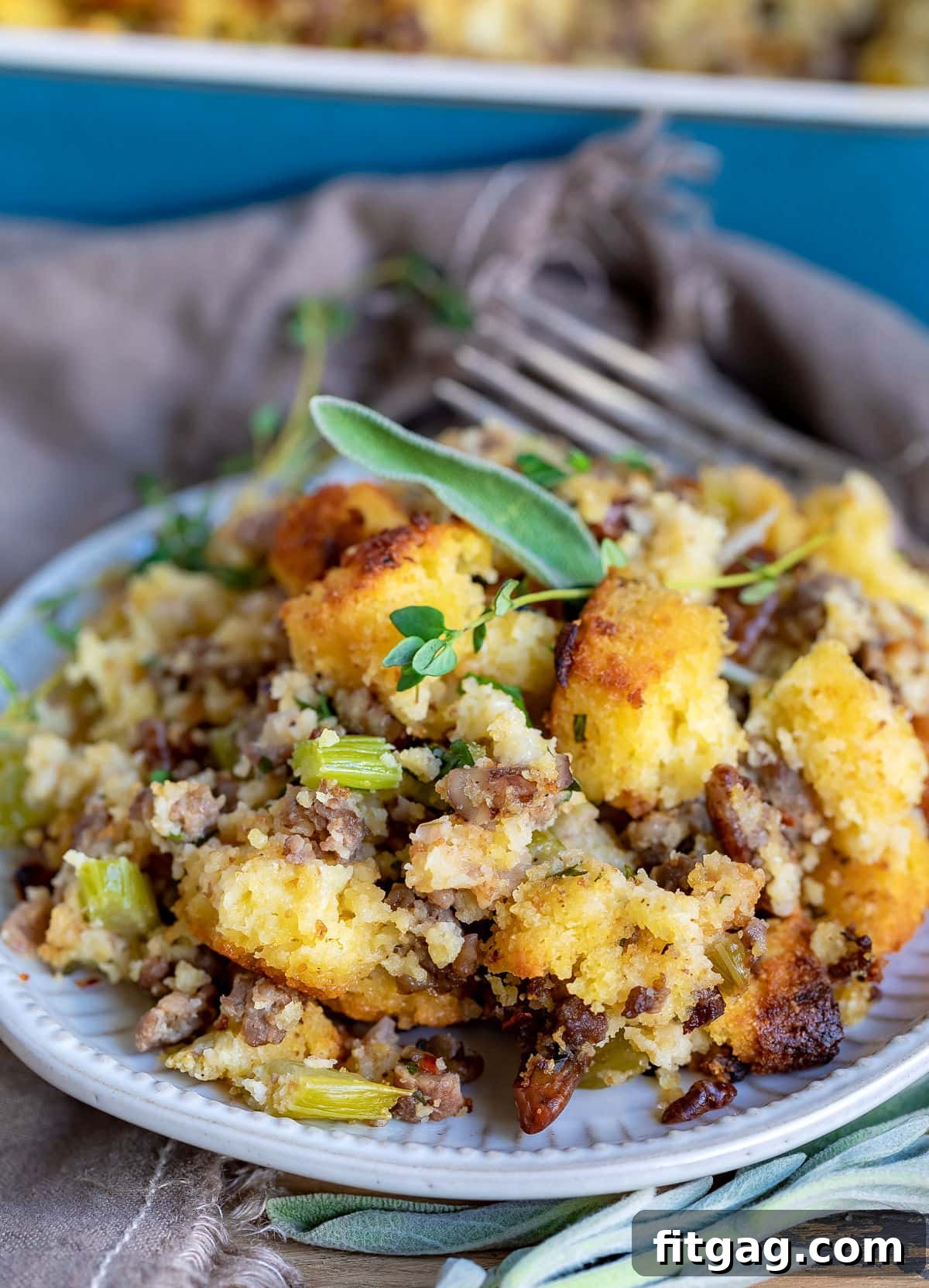 Ultimate Cornbread Dressing 9 A serving of golden-brown cornbread dressing on a white plate, garnished with a sprig of fresh herbs and a fork ready for tasting. This homemade cornbread stuffing is a buttery, savory, and moist Thanksgiving side dish, perfect for holiday meals.