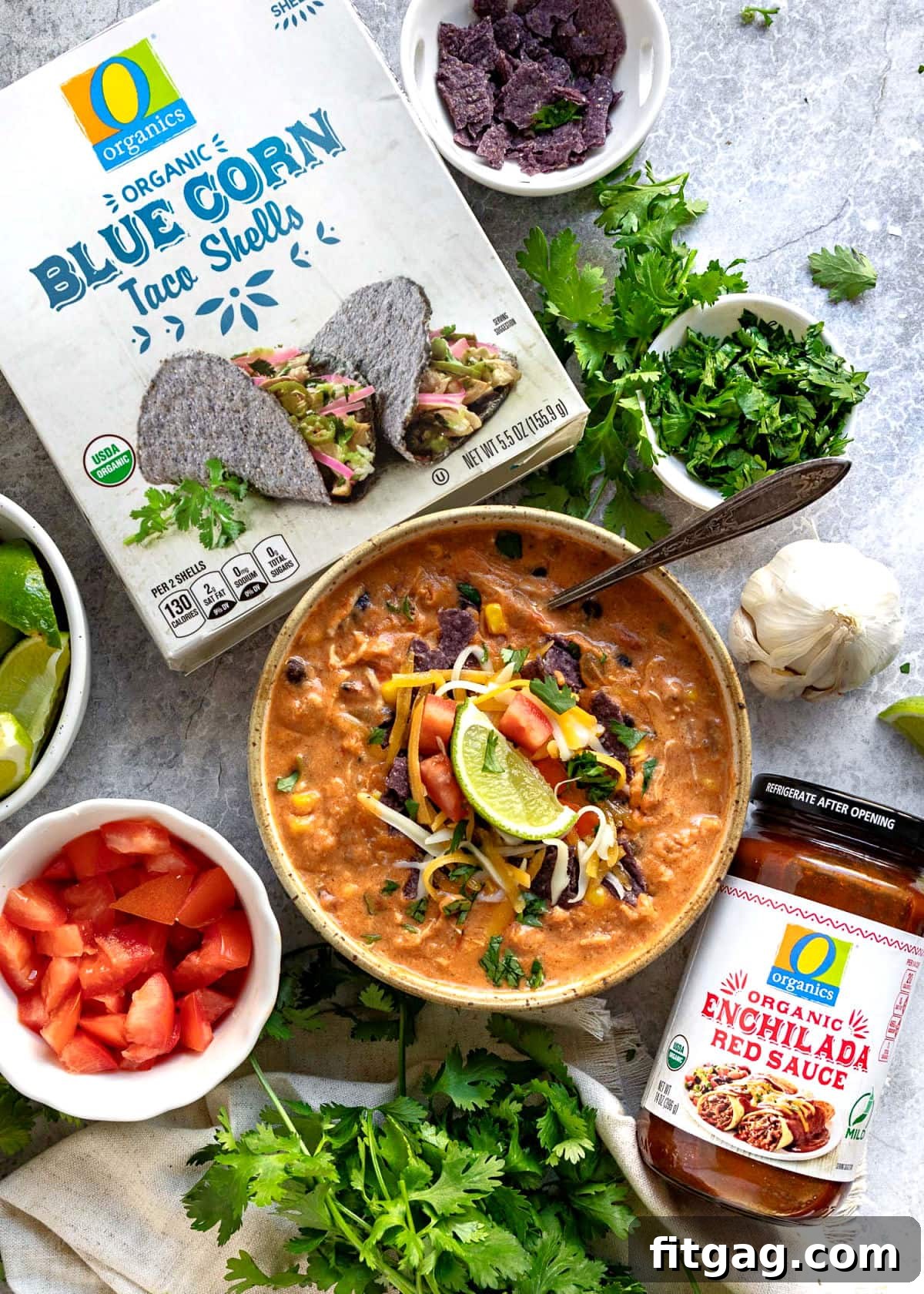 A top-down view of chicken enchilada soup in a bowl, surrounded by various topping ingredients in smaller bowls, alongside O Organics Enchilada Sauce and chicken broth packaging.