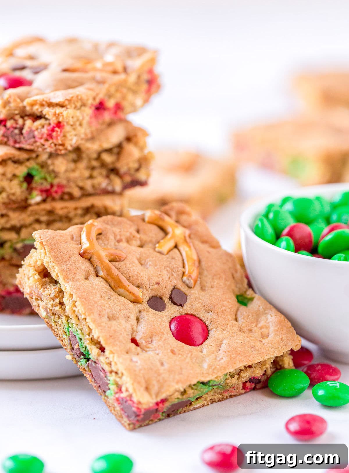 Magical Reindeer Cookie Bars 7 A single Reindeer Cookie Bar artfully leaned against a white plate, with a stack of more bars and a bowl brimming with red and green M&Ms, evoking a festive holiday scene.