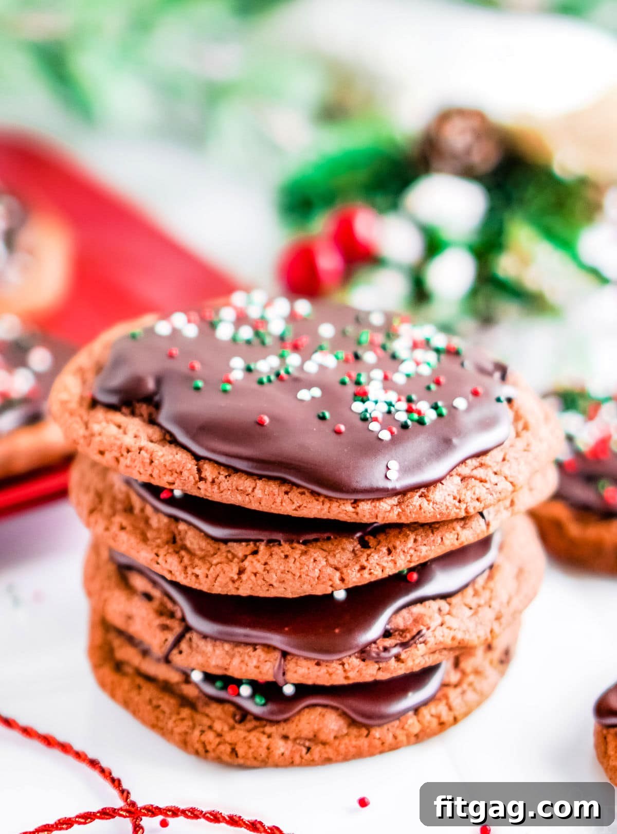 A stack of four perfectly glazed chocolate cookies, adorned with cheerful Christmas sprinkles, with festive holiday decor softly blurred in the background.